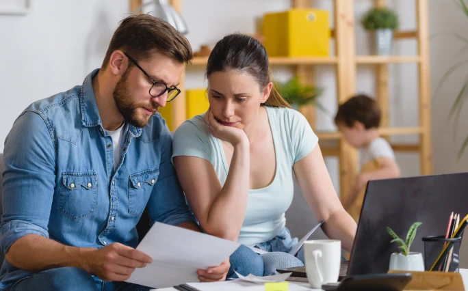 A puzzled man and woman are sitting at a table with a white cup and a black laptop. They both looking at a sheet of paper the man is holding, while their young son stands in the blurred background in front of wooden shelves
